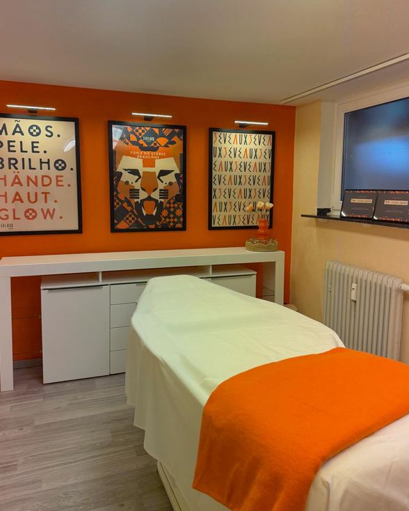 Behandlungsraum bei Shape and Flow: Massageliege mit orangefarbenem Plaid vor der orangefarbenen Wand mit Jeveaux-Postern