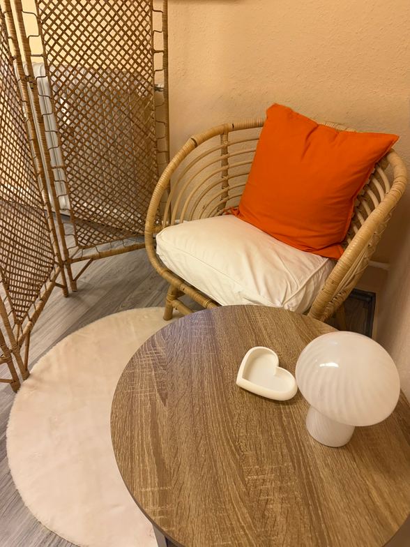 Umkleideecke im Studio mit Rattan-Paravent, Rattansessel mit orangefarbenem Kissen und kleinem Beistelltisch